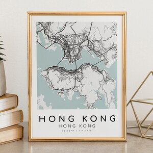 May include: A framed map print of Hong Kong, featuring a detailed black and white city plan against a light blue background. The text "HONG KONG" is displayed in a bold, sans-serif font, with coordinates below. The print is in a gold frame.