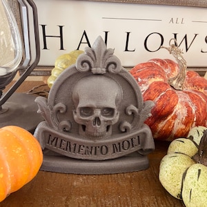 May include: A gray, stone-like decorative plaque featuring a central human skull relief set within a circular frame topped with a fleur-de-lis. A banner across the bottom is inscribed with the text MEMENTO MORI in raised capital letters.