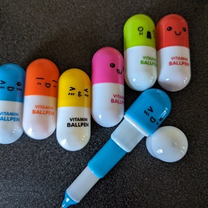 May include: A set of colorful pill-shaped ballpoint pens with different facial expressions. Each pen has the text "VITAMIN BALLPEN" printed on it.