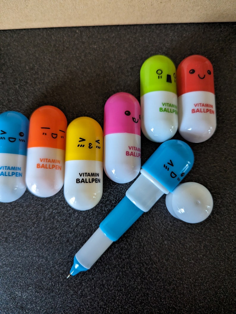 May include: A set of colorful pill-shaped ballpoint pens with different facial expressions. Each pen has the text "VITAMIN BALLPEN" printed on it.