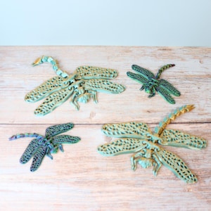 May include: Four decorative dragonfly sculptures in shades of blue, green, and gold are arranged on a light wood surface. The dragonflies have detailed wing patterns and are of varying sizes, creating a whimsical display.