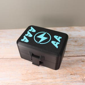 Könnte beinhalten: Eine schwarze Batteriestaufach mit einem Klappdeckel. Auf der Oberseite der Box steht in Türkis der Text "AAA" und "AA" sowie ein türkisfarbenes Blitzsymbol in einem Kreis. Die Box steht auf einer hellen Holzoberfläche.
