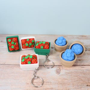 Puede incluir: Una colección de llaveros con cestas de frutas en miniatura. Los llaveros incluyen fresas rojas con tapas verdes en cestas blancas y verdes, y arándanos azules en cestas color canela. Los llaveros están sobre una superficie de madera.