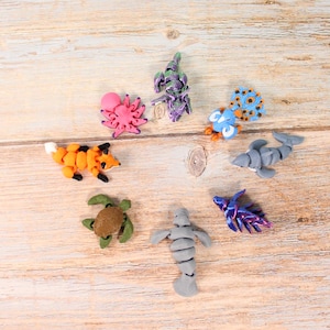 May include: A selection of handmade clay animal figurines, including a fox, turtle, shark, and manatee, displayed on a wooden surface. The figurines are brightly coloured and detailed, representing various sea and land creatures.