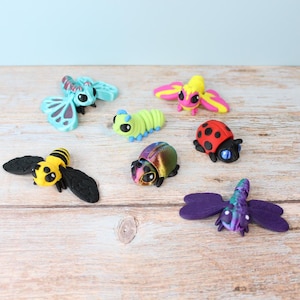 May include: A selection of brightly coloured insect figurines, including a ladybug, bee, butterfly, and caterpillar. The figurines are made from various materials and feature detailed designs. The insects are arranged on a wooden surface.
