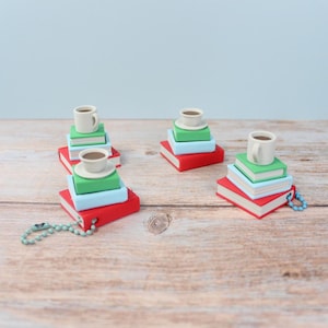 May include: Miniature stacks of books in red, light blue, and green, each topped with a tiny white teacup. Some stacks have a keychain attached. The background is a light blue wall and a wooden surface.
