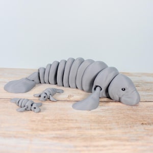 Puede incluir: Un juguete de manatí gris articulado con dos figuras de manatí más pequeñas. El manatí grande tiene un cuerpo segmentado y aletas. Los juguetes están sobre una superficie de madera clara.