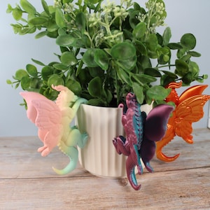Puede incluir: Varias figuras de hadas coloridas con alas extendidas, sujetas a una maceta de cerámica blanca. Las hadas son en tonos rosa, amarillo, azul y naranja. La maceta contiene una planta verde, sobre una superficie de madera clara.