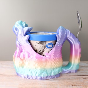 Op de afbeelding: Een veelkleurige ijspint houder met een lepel. De houder is een pastel regenboog van roze, paars, blauw en geel. De ijscontainer is blauw en wit met de woorden "Great Value". Een zilveren lepel zit in de houder.