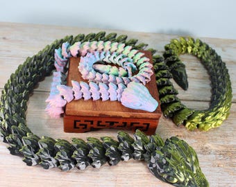 May include: Two colorful, segmented, snake-like sculptures. One is iridescent pink and green, the other is black and green. Both are curled up on a wooden box.