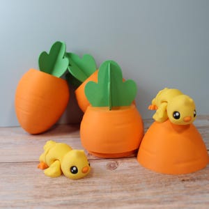 Pode incluir: Recipientes laranja em forma de cenoura com detalhes de folhas verdes, ao lado de pequenas figuras de pintinhos amarelos. Os recipientes são dispostos sobre uma superfície de madeira, criando uma exibição lúdica com tema de primavera.