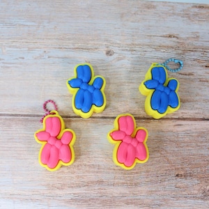 Puede incluir: Cuatro llaveros de perro globo coloridos. Dos son azules y dos rosas, cada uno con un contorno amarillo. Los llaveros están sobre una superficie de madera marrón claro. Cada llavero tiene una pequeña cadena de metal.