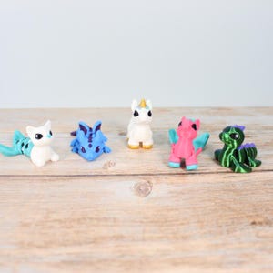 May include: A selection of five miniature fantasy figurines. The collection includes a white cat-like creature with a turquoise tail, a blue dragon, a white unicorn with a gold horn, a pink winged creature, and a green dragon.