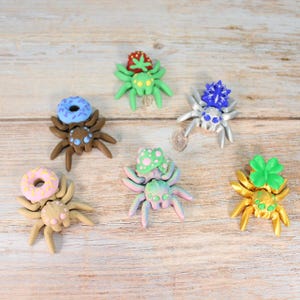 Op de afbeelding: Een verzameling van zes kleurrijke, grillige spinnenbeeldjes. Elke spin heeft een uniek ontwerp, waaronder een donut, een aardbei en een klavertjevier. De spinnen zijn gemaakt van klei en staan op een licht houten oppervlak.