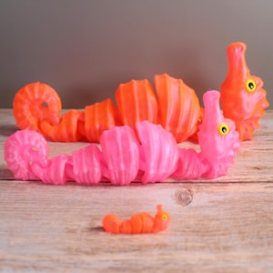 Può includere: Una collezione di giocattoli colorati e segmentati a forma di creature marine, nei toni dell'arancione e del rosa. I giocattoli sono disposti su una superficie di legno, con uno sfondo neutro. I giocattoli hanno un design stravagante e fumettistico.