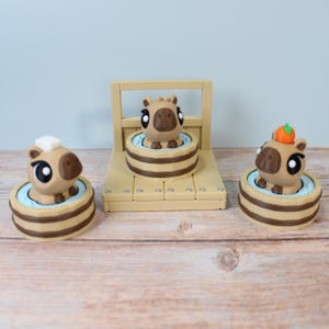 Puede incluir: Tres figuras de capibaras marrones y beige en bañeras de madera en miniatura. Una tiene un cuadrado blanco en la cabeza, otra una fruta naranja y la tercera está en una estructura de madera. Las bañeras tienen un interior azul claro.