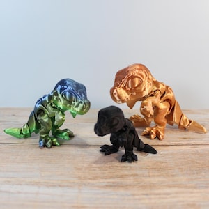 Könnte beinhalten: Drei 3D-gedruckte Dinosaurierfiguren in Grün-Blau, Gold und Schwarz. Die Spielzeuge sind in einer spielerischen Haltung dargestellt, mit detaillierten Texturen und Merkmalen. Sie werden auf einer Holzoberfläche präsentiert.