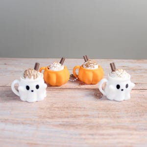 Könnte beinhalten: Vier Miniatur-Tassen zum Thema Halloween. Zwei sind wie weiße Geister geformt, zwei wie orangefarbene Kürbisse. Jede Tasse ist mit weißer Schlagsahne, Schokoladensauce und einem Schokostäbchen gefüllt. Die Tassen stehen auf einer Holzoberfläche.