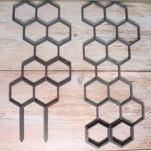 May include: Two gray, plastic plant trellis panels with a honeycomb design. Each panel has a series of connected hexagons, with stakes at the bottom for ground insertion. The panels are designed for supporting climbing plants.