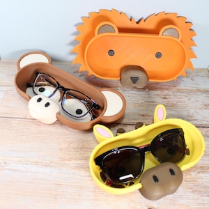 May include: Three whimsical eyeglass holders in the shape of animals. The top holder is orange and shaped like a lion. The middle holder is brown and shaped like a bear. The bottom holder is yellow and shaped like a giraffe. Each holder contains a pair of eyeglasses or sunglasses.