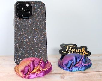 May include: A black phone case with a colourful paint splatter design stands next to two metallic triceratops phone holders. One holder is pink and gold, the other is blue and purple, holding a black sign that says "Thank You".