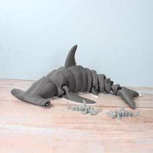 Puede incluir: Una figura articulada grande de tiburón martillo en color gris con un diseño de cuerpo segmentado, acompañada por dos figuras de tiburón martillo grises más pequeñas, todas con superficies lisas y cabezas en forma de martillo.