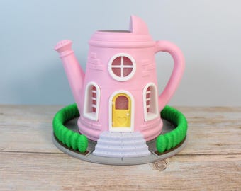 May include: A pink watering can shaped like a house with a yellow door and white windows. The house is surrounded by a green hedge.
