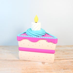 May include: A pink and white cake slice with blue frosting and a single yellow candle on top. The cake is decorated with small blue dots around the edges.