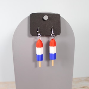 Può includere: Un paio di orecchini a forma di ghiacciolo rosso, bianco e blu. Gli orecchini sono appesi a ganci d'argento e hanno un bastoncino di legno. Sono esposti su un espositore per gioielli grigio.