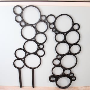 May include: Two black, laser-cut wooden plant stakes with a bubble design. Each stake features a series of interconnected circles of varying sizes, creating an abstract, decorative element. The stakes are designed to be inserted into soil.