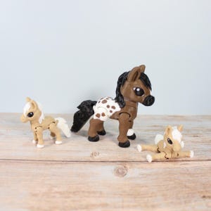 Könnte beinhalten: Drei Spielzeugpferde werden auf einer Holzoberfläche gezeigt. Das mittlere Pferd ist braun mit weißen Flecken und einer schwarzen Mähne und einem schwarzen Schweif. Die anderen beiden Pferde sind hellbraun mit weißen Mähnen und Schweifen. Die Pferde sind beweglich.