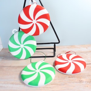 Può includere: Quattro decorazioni natalizie rotonde con un design a caramella alla menta piperita. Due sono rosse e bianche, e due sono verdi e bianche. Le decorazioni sono appese a un espositore nero a forma di albero.