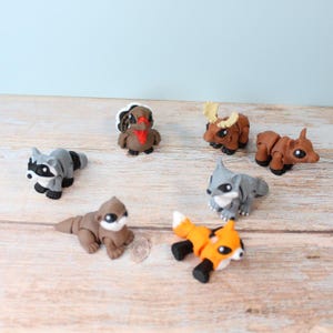 Puede incluir: Colección de figuras de animales hechas a mano, incluyendo un mapache, un pavo, un alce, un oso, un lobo, un zorro y una nutria. Las figuras son de arcilla y presentan detalles en las facciones y el color. Los animales están dispuestos sobre una superficie de madera.