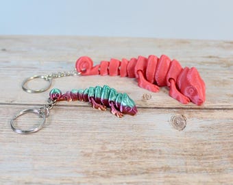 Könnte beinhalten: Zwei bewegliche Chamäleon-Schlüsselanhänger. Einer ist in mattem Rot gehalten, der andere zeigt einen Farbverlauf in metallischem Grün und Violett. Beide Figuren haben segmentierte Körper, die sich bewegen lassen, und sind an silbernen Metallringen befestigt.