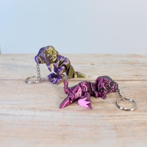 Op de afbeelding: Twee kleine dinosaurus sleutelhangers. Eén is paars en goud, de andere is roze en paars. Beide hebben zilveren sleutelringen.