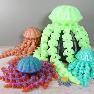 May include: A collection of colorful, 3D-printed jellyfish sculptures in various sizes. The jellyfish feature detailed tentacles and bell-shaped bodies in shades of coral, blue, green, and purple. The sculptures are arranged on a light-colored surface.