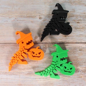 Könnte beinhalten: Drei Halloween-Dinosaurierfiguren in Orange, Schwarz und Grün. Jeder Dinosaurier hält einen Kürbis. Die Figuren bestehen aus einem segmentierten Material und tragen jeweils einen Hexenhut.