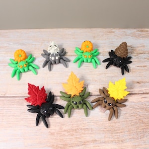 May include: A selection of handmade polymer clay spiders in various colours. Each spider features unique details, including pumpkins, skulls, and leaves. The spiders are arranged on a light wood-grain surface, ideal for Halloween decor.