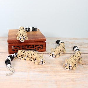 May include: Several small, tan cheetah figurines with black spots and striped tails. Some are lying down, others are on a small wooden box. The cheetahs are on a light-coloured wooden surface.