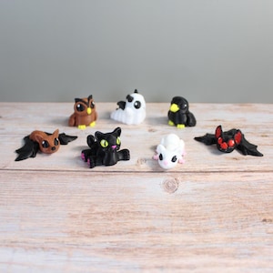 May include: A collection of small, whimsical figurines of various creatures, including bats, an owl, a cat, and a ghost. The figurines are in shades of brown, black, white, and pink, with yellow accents. They are arranged on a light-colored wooden surface.
