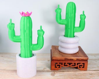 May include: Two green cactus figurines with raised middle fingers, one with a pink flower on top, both in white pots. The cactus on the right is on a wooden stand.