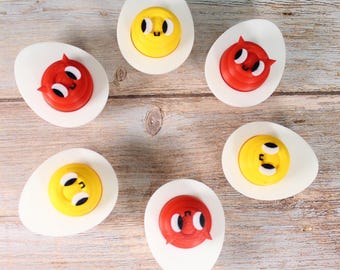 Pode incluir: Seis metades de ovos cozidos brancos dispostas em círculo, cada uma coberta com uma gema colorida com rosto de desenho animado. As gemas são vermelhas ou amarelas, com olhos pretos e uma boca sorridente. Os ovos estão sobre uma superfície de madeira castanha clara.