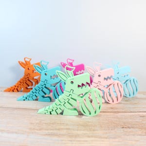 May include: Five colorful dinosaur-shaped doorstops in orange, blue, green, pink, and light blue. Each doorstop has a textured surface and a cartoonish dinosaur design. The doorstops are arranged in a row on a wooden surface.