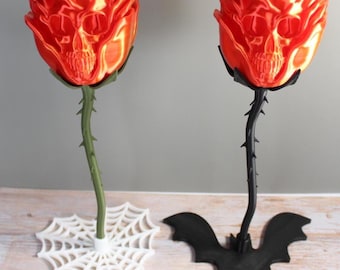 May include: Two decorative sculptures of red roses with integrated skull faces. One rose has a green stem on a white spiderweb base, and the other has a black stem on a black bat-shaped base. Both items feature thorny stems and glossy petals.