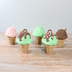 Può includere: Una collezione di cinque ornamenti a forma di cono gelato in miniatura. I coni sono di colore marrone chiaro e le palline di gelato sono rosa, verdi e marroni, con glassa al cioccolato e zuccherini. Gli ornamenti sono esposti su una superficie di legno.