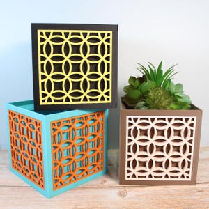 Op de afbeelding: Drie vierkante plantenbakken met geometrische uitsparingen. De plantenbakken zijn zwart, turquoise en bruin. De uitsparingen zijn geel, oranje en crème.