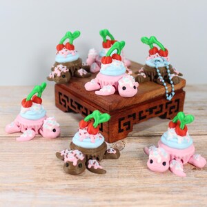 以下が含まれることがあります： ピンク色の体と茶色の甲羅を持つ、デザートとさくらんぼがトッピングされた、気まぐれなカメの置物のコレクション。カメは木製の表面に配置されています。
