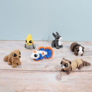 May include: A collection of six small animal-shaped erasers including a gray cockatiel, a brown dog, a white and blue betta fish, a gray rabbit, a brown and white guinea pig, and a brown ferret, all featuring large black eyes and detailed forms.