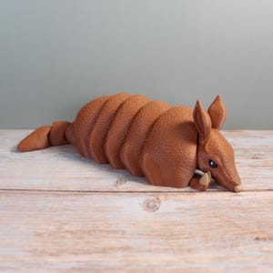 Peut inclure: Figurine d'un tatou brun avec une carapace texturée et des détails précis. Le tatou est allongé sur une surface en bois, mettant en valeur son corps segmenté et ses petites oreilles. Objet décoratif.