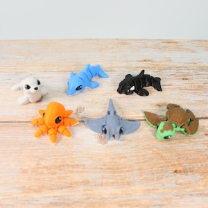 Puede incluir: Una colección de criaturas marinas de arcilla hechas a mano, que incluyen una foca blanca, un delfín azul, una orca negra, un pulpo naranja, una raya gris y una tortuga marina verde y marrón. Las figuras están dispuestas sobre una superficie de madera clara.
